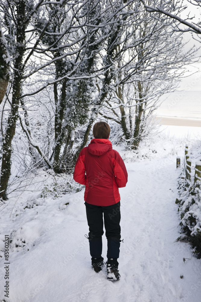 Red Wharf Bay (Traeth Coch), Isle of Anglesey, North Wales, UK, Europe.  Walker in red jacket on snow covered Isle of Anglesey Coastal Path leading to the beach in winter. 