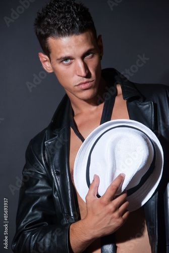 Fashionable topless male with white fedora hat, leather jacket and tie, studio shooting black background