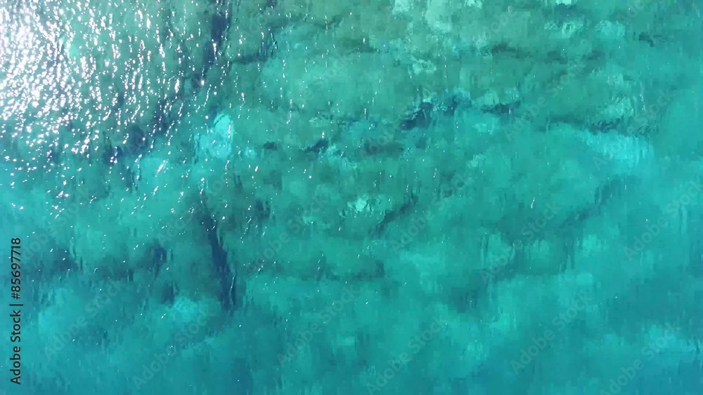 Escena con dron en planta del agua en una bahía. Agua turquesa. Rocas aparecen debajo. Camara se acerca, hace zoom. Detalle. Textura de agua en movimiento.