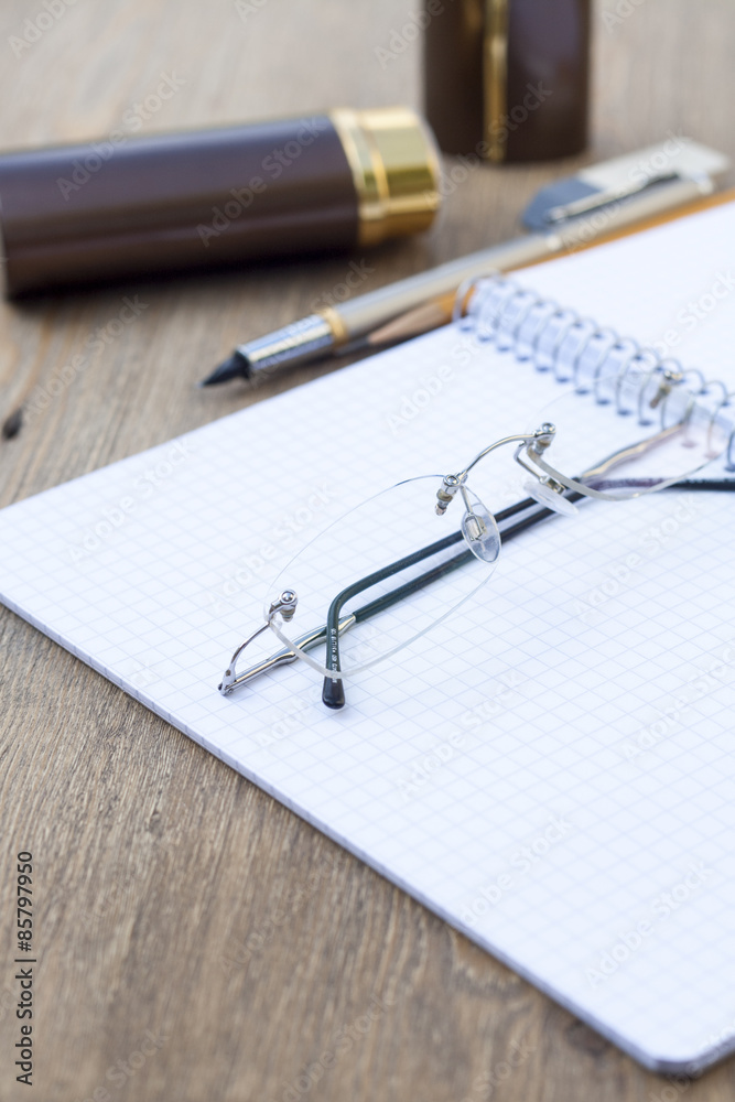 Expand spiral notebook into a cell with a pen, pencil, eraser and glasses on a wooden table, selective focus Expand spiral notebook into a cell with a pen, pencil, eraser and glasses on a wooden table, selective focus