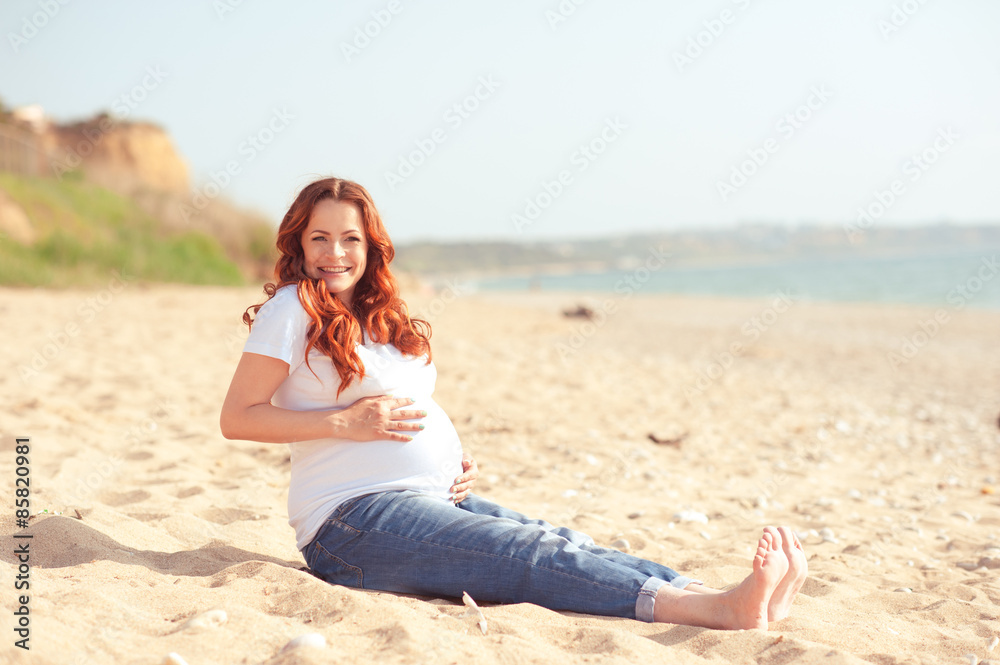 Smiling pregnant woman sitting on seashore. Looking at camera. Motherhood. Maternity.