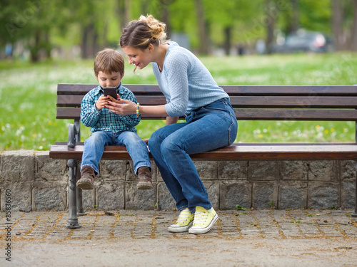 Cute little boy sitting on a park bench with his mother and playing with mobile phone