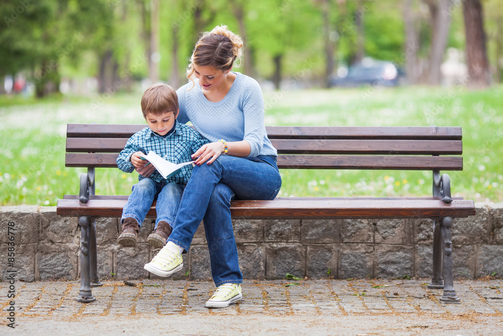 Mother and son sitting on a bench in a park and reading a book - obrazy, fototapety, plakaty 