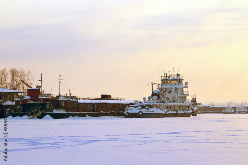 Fototapeta premium Little ship in frozen river covered with snow in winter evening