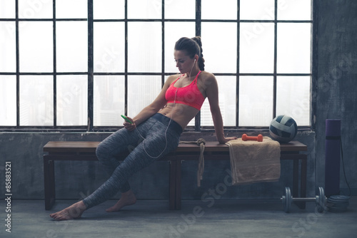 Fit woman in loft gym sitting on bench selecting music