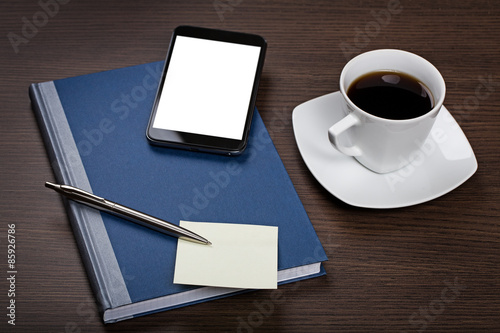 Top view on dark wooden office desk with notepad, pen, sticky note, mobile with blank screen and cup of coffee.