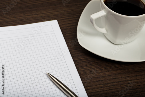 Close up on notepad, pen and cup of coffee on dark wooden office desk.