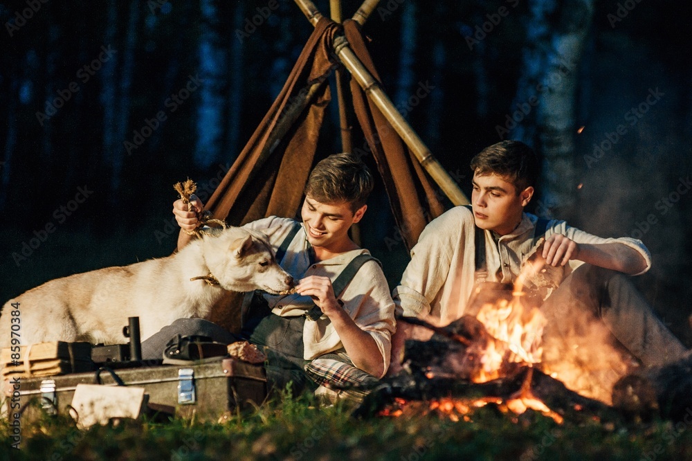 two twin brothers sitting around a campfire in a birch grove