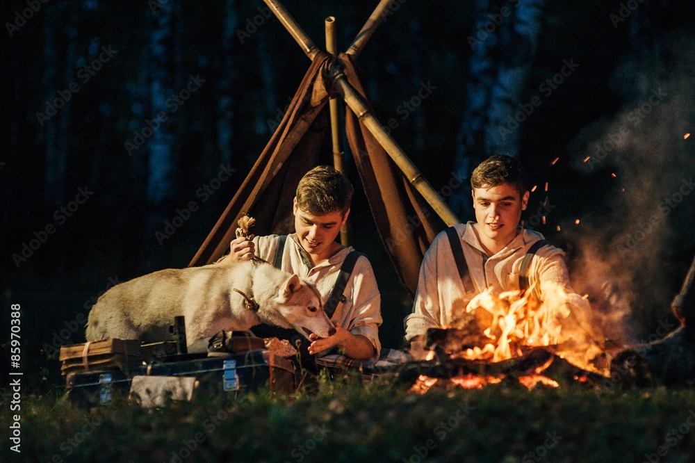 two twin brothers sitting around a campfire in a birch grove
