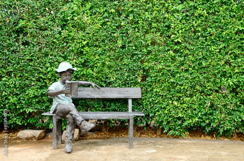 Boy reading on bench statue and green tree wall