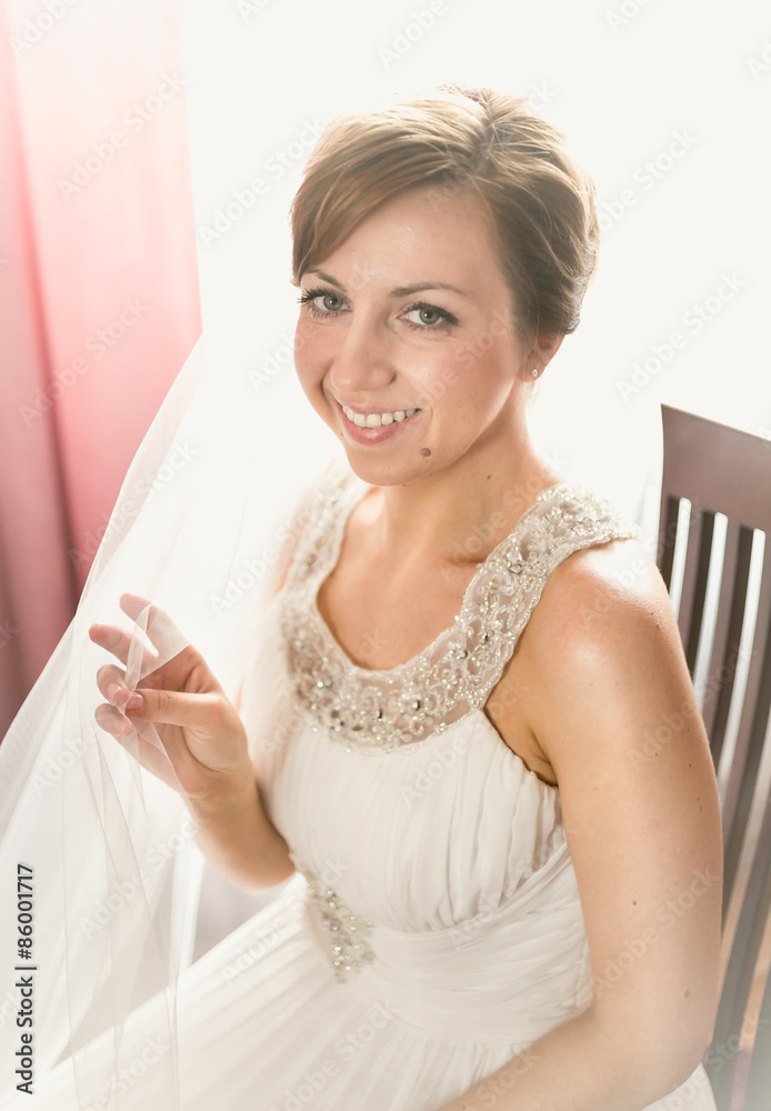smiling bride sitting on chair next to window at bedroom smiling bride sitting on chair next to window at bedroom