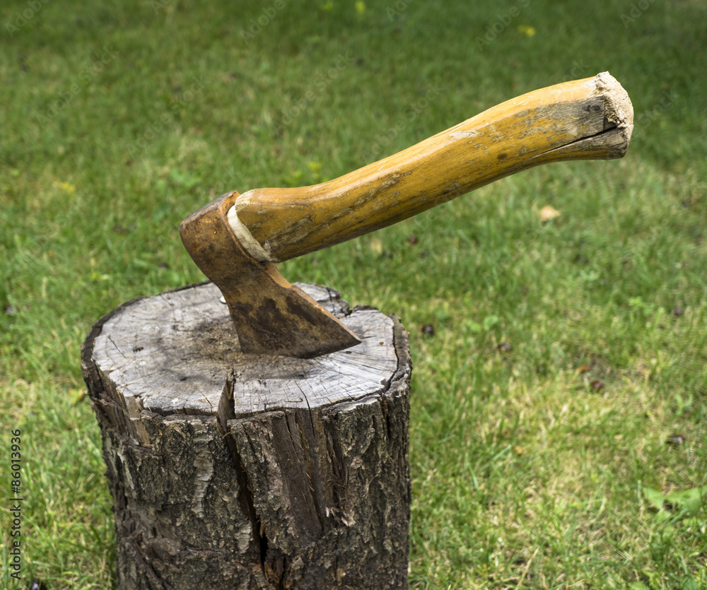 Ax driven into the wood timber in the yard on background of green grass Ax driven into the wood timber in the yard on background of green grass