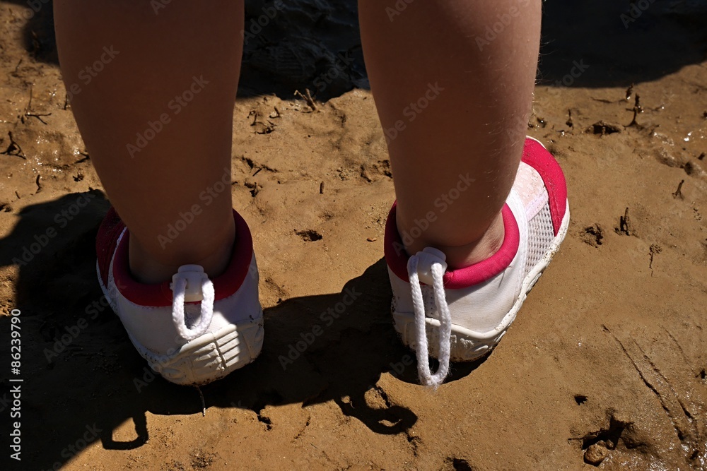 Small girl legs in protective aquatic shoes standing on muddy sandy beach Small girl legs in protective aquatic shoes standing on muddy sandy beach