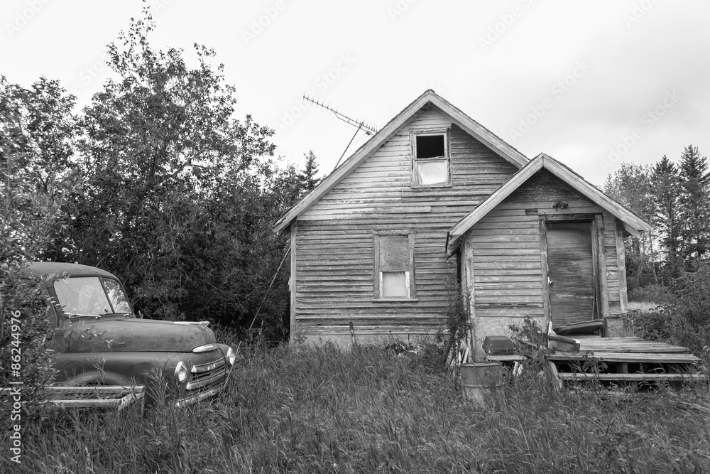 horizontal black and white image of an old abandoned house with a porch and an old vintage truck sitting in the yard in the summer time.