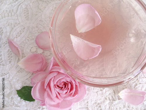 Kristallschale mit Rosenwasser gefüllt, daneben eine englische Rose und Rosenblätter, alles auf nostalgischem Deckchen.