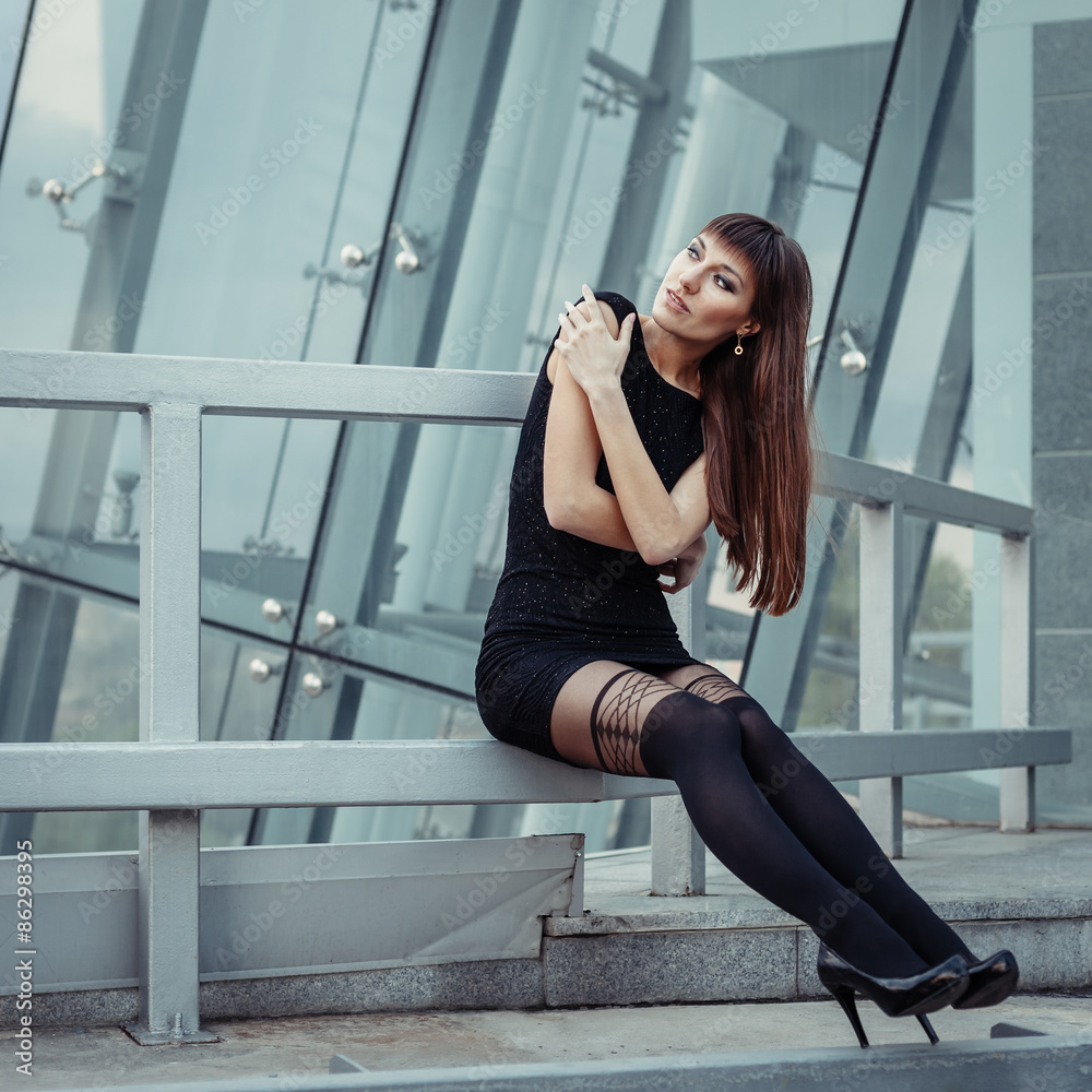 Fashion outdoor portrait of sexy beautiful woman with brown hair wearing in black dress, hose and high heels. Posing on creative urban background.