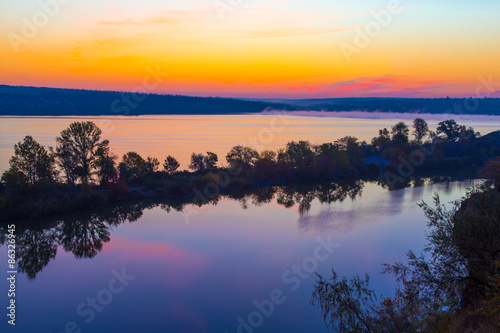 Amazing orange dawn on blue lake. Luminous sunrise blooming over wild waterfront landscape with fall grass flower hill stone rocks and narrow island with colored trees in the middle of water surface