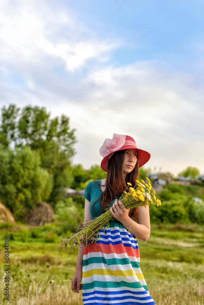 Portrait of a young female standing in field holding flowers wearing red hat