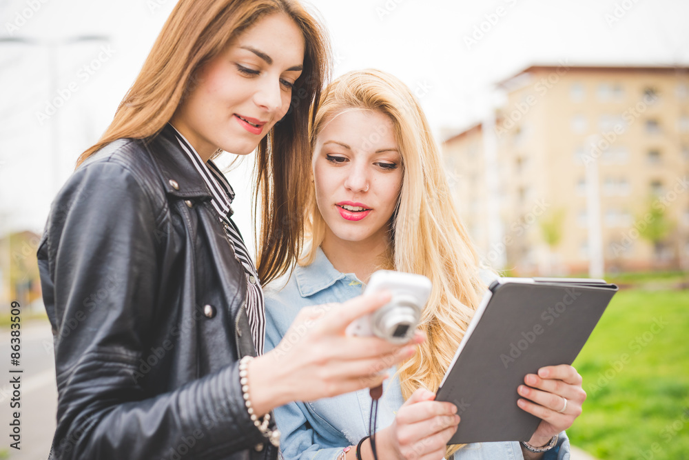 Two young blonde and brunette girls walking through the city using a tablet and a camera - technology, communication, social network concept Two young blonde and brunette girls walking through the city using a tablet and a camera - technology, communication, social network concept