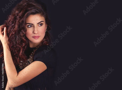 Beautiful young latin woman face, holding her long wavy hair, looking to the left and thinking on a black background with empty copy space