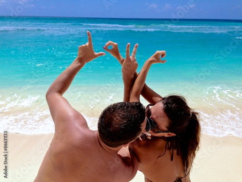 A couple kissing on a Caribbean beach, making the letters LOVE with their hands.