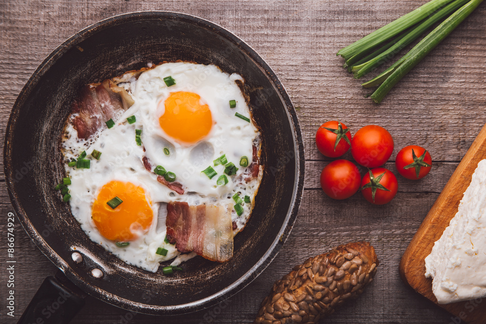 Two fried eggs with bacon and onion, tomato and bread on the side
Two fried eggs with bacon and onion, tomato and bread on the side