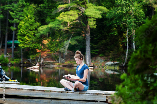 girl sitting on a dock by a lake reading a book © sianc
