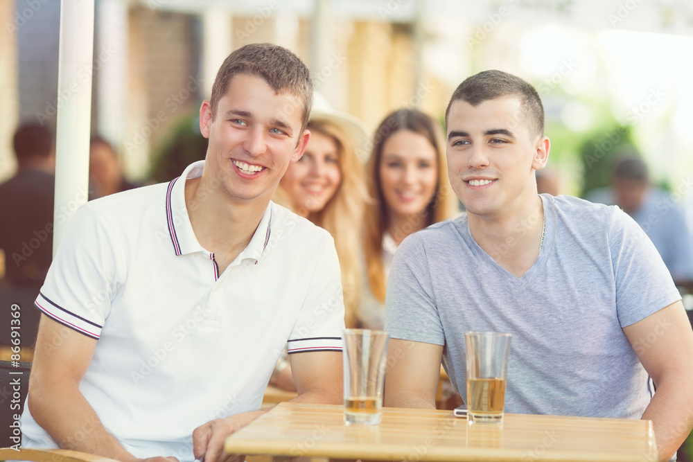 Two young friends sitting in a bar drinking beer and checking out girls passing by
