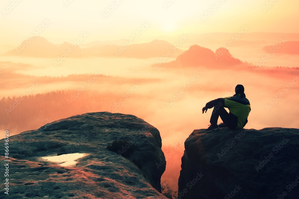 Rear view of male hiker sitting on the rocky peak  while enjoying a colorful daybreak above mounrains valley - obrazy, fototapety, plakaty 