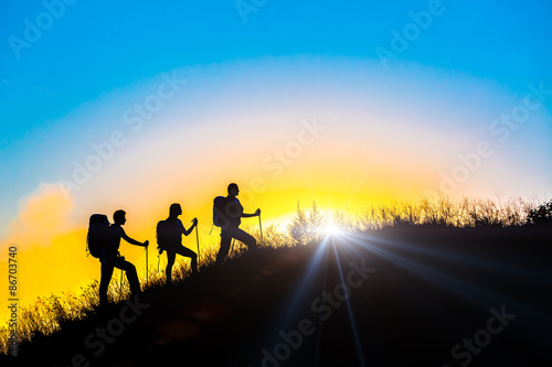 People meeting sunrise on team building session Group of people silhouettes walking toward mountain summit with backpacks hiking trekking gear meeting uprising sun sunbeams and blue sky of background © alexbrylovhk