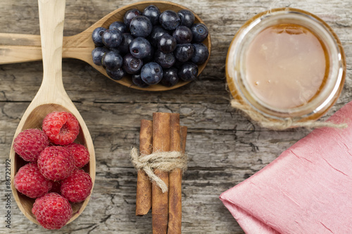 Raspberries and blueberries in spoons with pink napkin and a jar of honey with cinnamon on wooden background. Healthy eating, diet