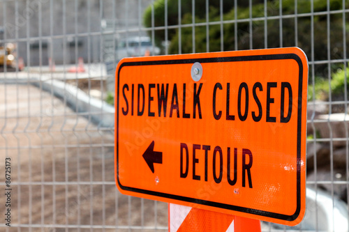 Orange sidewalk closed detour sign in front of fence and construction