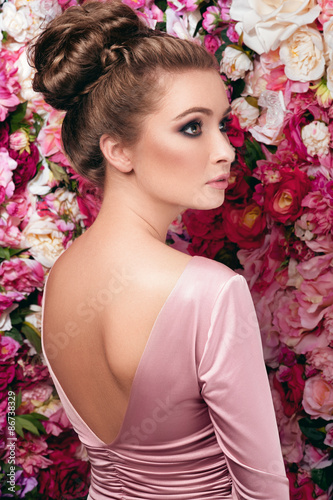 Fototapeta Naklejka Na Ścianę i Meble -  Soft facial portrait of young charming woman in rosy skin-tight gown with V-shaped cut and open back, dark smoky eyes and natural lipstick standing behind on flowered background made of peony
