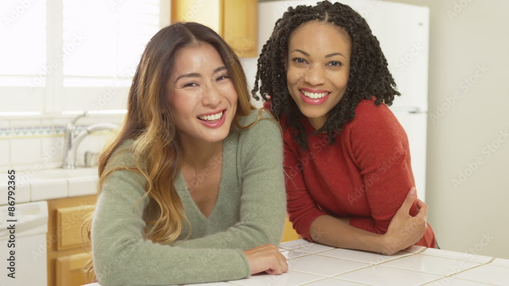 Black and Japanese friends leaning against kitchen counter smiling Black and Japanese friends leaning against kitchen counter smiling