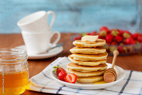 Stack of pancakes with honey syrup, butter and strawberry in a white plate on a wooden rustic table, selective focus