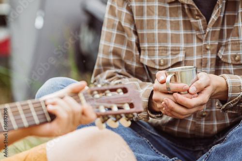 A person holding a cup of tea while his friend plays the guitar