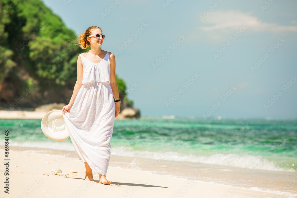 Young woman in long white dress holding hat walks along tropical Young woman in long white dress holding hat walks along tropical