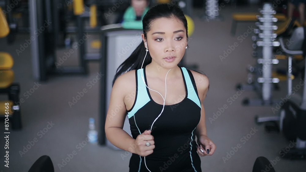 Attractive asian girl running on the treadmill in the gym with earphones. Full face portrait