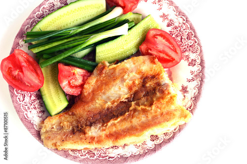 Кусок жаренной рыбы, лук, помидоры, огурцы на тарелке на белом фоне