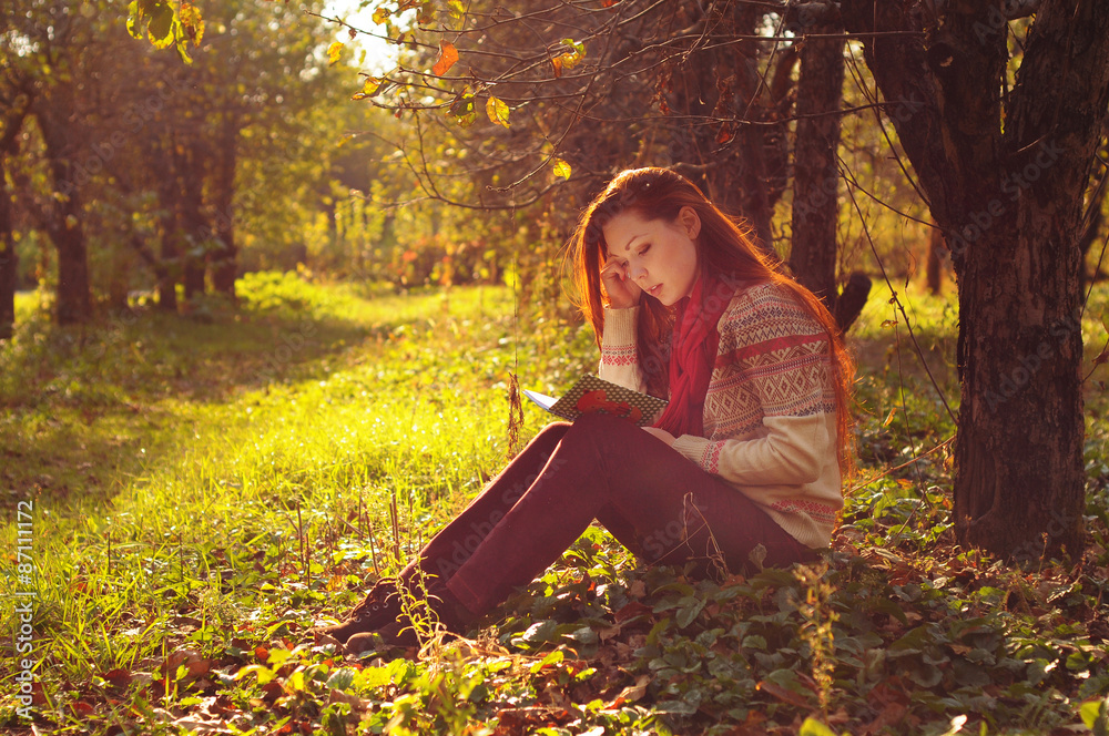 Young woman with long red hair reading under the tree