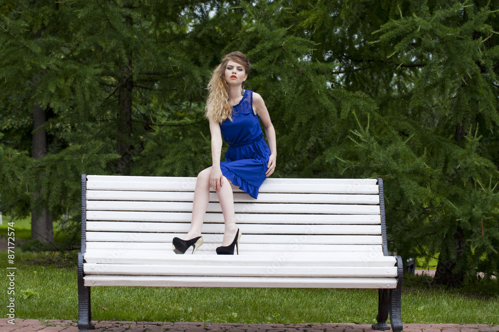 Beautiful blonde girl in blue dress sitting on a bench in summer Beautiful blonde girl in blue dress sitting on a bench in summer