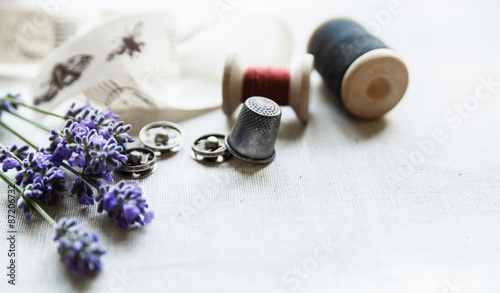 Sewing tools with fresh lavander flowers on linen background. Vintage wooden spool, braid, thimble, buttons.