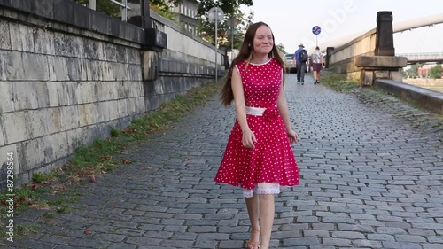 Young pretty girl in a red dress goes merrily skipping along pavement city waterfront