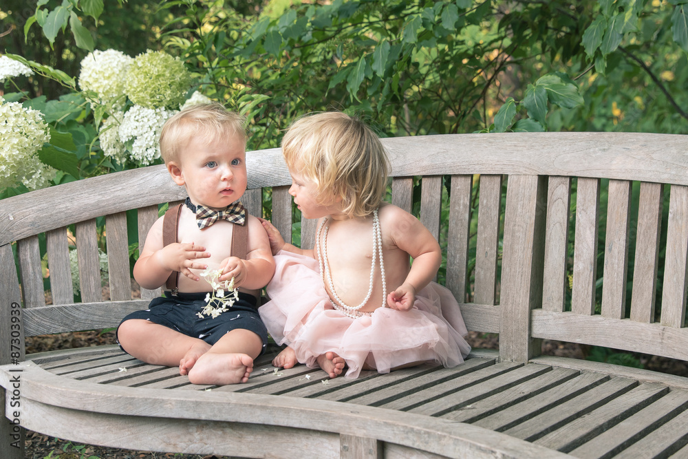 Fototapeta premium Baby boy and girl in formal dress sitting on wooden bench in a beautiful garden
