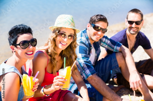 Vier Freunde sitzen mit Getränken am See Strand in der Sommersonne