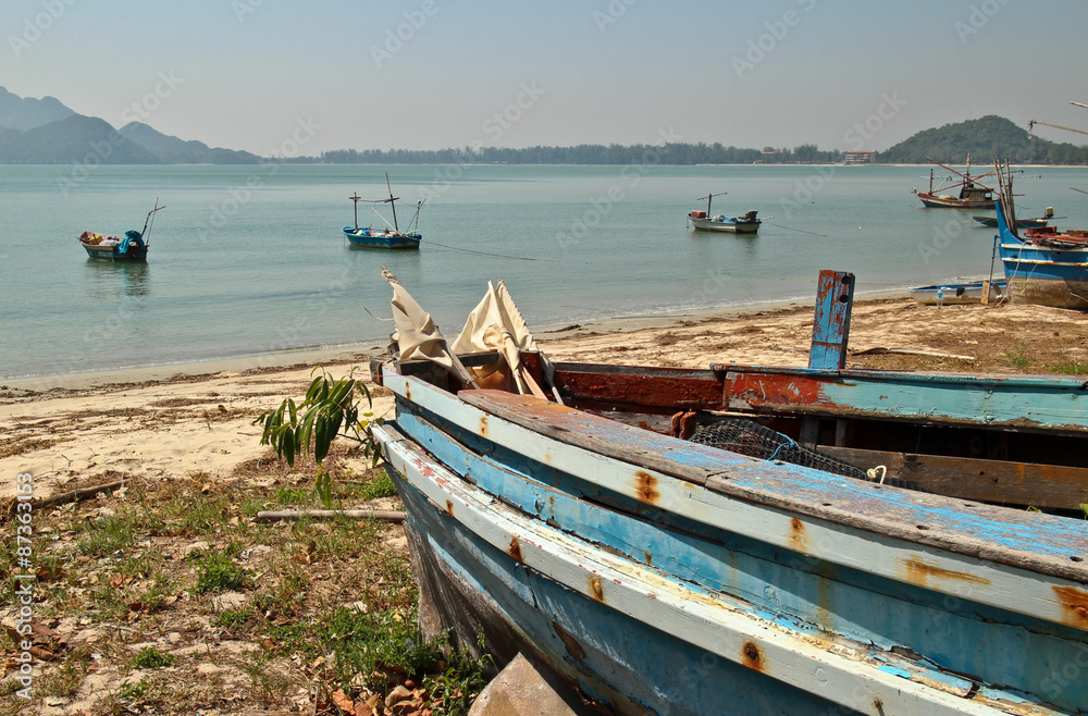 Weathered wooden vintage fishing boat on shore at a calm bay in the sea along the Southern Coast of Thailand; several other boats are visible just offshore, with mountains in the distance