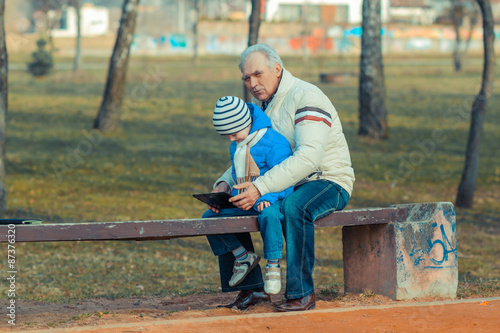 Grandfather and grandson are looking tablet on bench outdoors