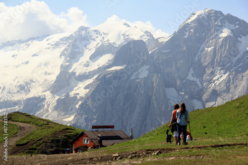 Woman and girl hiking in the Dolomites. Marmolada in Background. Hikers on the path with green Grass, vertical rockwalls and white glacier in background