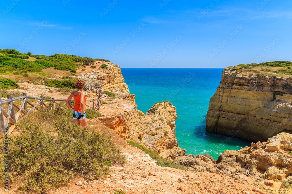 Young woman tourist standing on cliff rock and looking at sea on coast of Portugal, Algarve region
