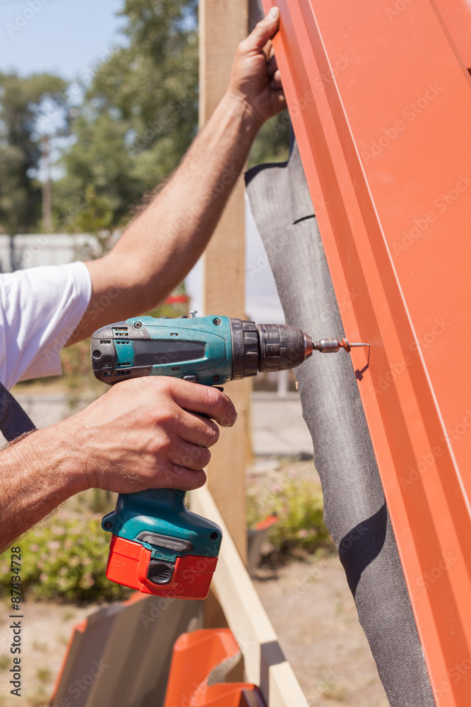 Worker on a roof with electric drill installing red metal tile on wooden house Worker on a roof with electric drill installing red metal tile on wooden house